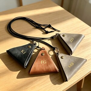 May include: Four triangular pouches in black, tan, and gray, each featuring a gold-toned snap button and gold-embossed initials KJ, FG, ZD, and MB. Each pouch is attached to a thin black cord with a metal ring, designed for carrying small items.