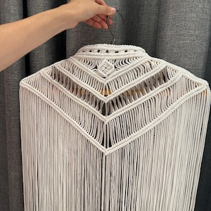 May include: A white macrame wall hanging with a geometric design. The piece features a detailed woven pattern at the top, with long, vertical fringe cascading downwards. The handmade textile is displayed on a wooden hanger, showcasing its texture and craftsmanship.