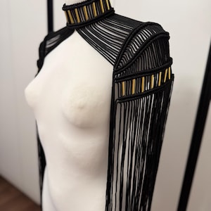 May include: A black macrame shoulder piece featuring a high neck collar and long fringe detailing. The design incorporates vertical gold-toned metal tubes integrated into the black cord structure, extending from the neck down to the shoulders.