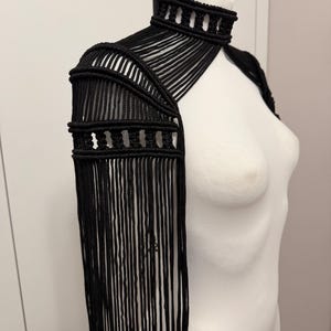 May include: A black macrame shoulder piece featuring a high neck collar, intricate knotted patterns, and long, flowing fringe that extends down the arm. The garment is constructed from cord and displays a structured, layered design on the shoulder.
