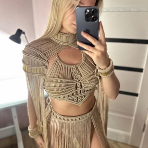 May include: A beige macrame two-piece set featuring a high-neck crop top with intricate knotted patterns and gold-toned accents, paired with a matching floor-length skirt with long fringe detailing and a high side slit, plus two matching wrist cuffs.