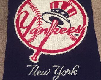 May include: A blue knitted blanket with a white and red New York Yankees logo. The logo features a baseball, a bat, and the team's name. The words "New York" are written in white below the logo.
