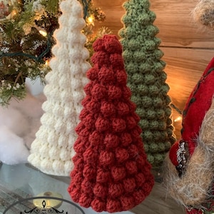 May include: Three handmade crochet Christmas trees in white, red, and green. The trees have a textured, bobble stitch design. The trees are displayed on a glass surface with Christmas decorations in the background.