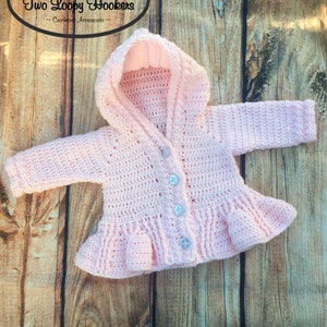 May include: A pink crocheted baby cardigan with a hood and buttons. The cardigan has a ruffled hem and is made with a delicate stitch pattern.