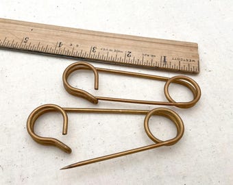 May include: Three gold safety pins, each with a different size and shape of the pin head. The pins are on a white surface.