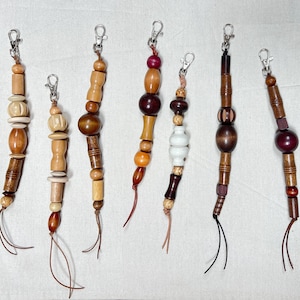 May include: A collection of seven wooden bead keychains with silver-tone clasps. The beads vary in shape, size, and color, ranging from light to dark brown, with some featuring white or reddish accents. Each keychain has a leather cord.