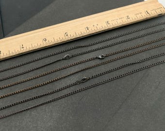 May include: Three dark gunmetal curb chains with lobster claw clasps, positioned alongside a wooden ruler that shows the chains measure approximately 8 inches in length. The metal links are uniform in size and have a smooth, metallic finish.