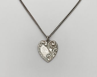 May include: A silver necklace with a heart-shaped pendant. The pendant has a detailed, ornate design with swirls and leaves.