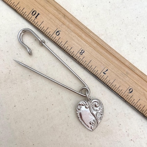 May include: A silver safety pin with a decorative heart charm. The heart charm has a swirling design.