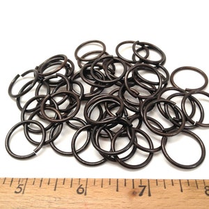 May include: A collection of dark brown metal jump rings scattered above a wooden ruler. The ruler shows measurements in inches, with the rings ranging in size and featuring a small opening on each circular band for connecting jewelry components.