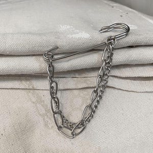 May include: A silver-colored safety pin with two attached chains. The pin is open and the chains hang down. The background is a white, textured fabric.