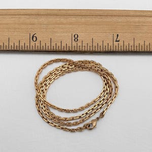 May include: A gold-colored chain necklace with a rectangular link design, approximately 18 inches long, is coiled on a white surface. A wooden ruler with inch markings is in the background.