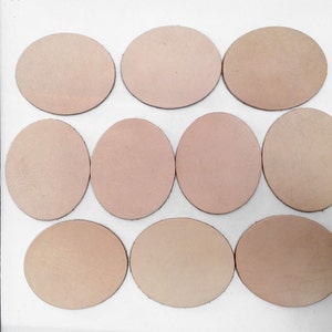May include: Twelve oval-shaped pieces of light brown leather are arranged on a white surface. A wooden ruler is positioned vertically along the left side. The leather pieces vary slightly in color, with some appearing more pinkish.