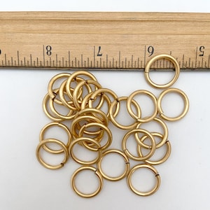 May include: A collection of gold-colored jump rings, some open, scattered on a white surface. A wooden ruler with inch markings is partially visible in the background. These rings are likely used for jewelry making or crafts.