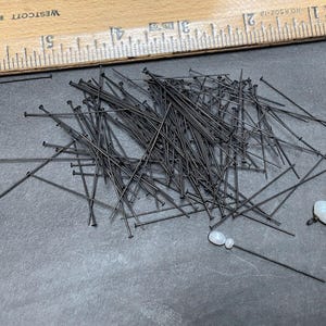 May include: A large quantity of black straight pins scattered on a gray surface. Two white oval-shaped stones are also visible.