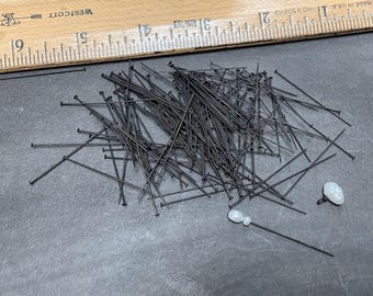 May include: A large quantity of black straight pins scattered on a gray surface. Two white oval-shaped stones are also visible.