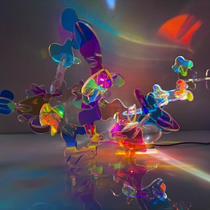 Puede incluir: Una escultura abstracta moderna con una compleja disposición de formas iridiscentes multicolores. La escultura está iluminada, proyectando patrones de luz coloridos sobre la superficie. La pieza tiene una estética futurista y artística. Medidas en centímetros.