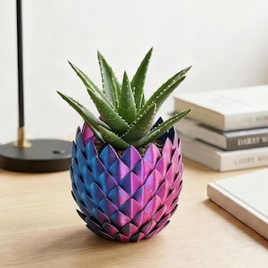 May include: A green aloe vera plant sits inside a decorative planter with a scale-like texture. The planter features a vibrant gradient finish that transitions from deep blue at the base to a bright magenta purple at the top rim.