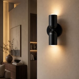 Könnte beinhalten: Moderne schwarze Wandleuchte mit zwei zylindrischen Leuchten. Die Leuchte ist auf einer kreisförmigen Basis montiert und strahlt warmes Licht aus, wodurch eine gemütliche Atmosphäre entsteht. Das Design ist minimalistisch und für verschiedene Einrichtungsstile geeignet.