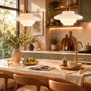 May include: Two white tiered glass pendant lights with brass hardware hang above a kitchen island. The lights feature a wide, multi-layered circular design that emits a soft glow, illuminating the marble countertop and wooden bar stools below.