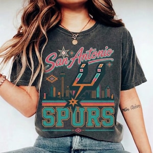 May include: A charcoal gray short-sleeve t-shirt featuring a graphic print with the text San Antonio SPURS. The design includes a stylized city skyline, the Spurs logo, and geometric patterns in shades of teal, orange, and pink.