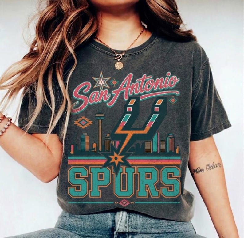 May include: A charcoal gray short-sleeve t-shirt featuring a graphic print with the text San Antonio SPURS. The design includes a stylized city skyline, the Spurs logo, and geometric patterns in shades of teal, orange, and pink.