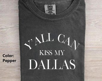 May include: A pepper-colored short-sleeve t-shirt with a crew neckline. The shirt features white text printed on the front that reads Y'ALL CAN KISS MY DALLAS. A Comfort Colors brand label is visible on the inside of the collar area.