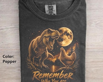 May include: A charcoal gray crew neck t-shirt featuring a graphic print of a Tyrannosaurus Rex and a chicken standing before a large full moon. The text "Remember Who You are." is printed in a golden-brown script font below the animal figures.