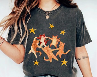 May include: A charcoal gray short-sleeve t-shirt featuring a graphic print of four orange cats holding hands in a circle, surrounded by several yellow stars. The cats are depicted in a stylized, artistic manner with visible brushstroke textures.