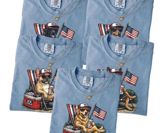 May include: Light blue Comfort Colors t-shirts featuring various dog breeds wearing sunglasses and patriotic hats, holding American flags and drinks. Each dog is depicted relaxing in a beach chair with an "Ice Cold" cooler. Gold chain necklaces are also visible.