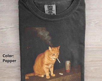 May include: A charcoal gray t-shirt featuring a graphic print of an orange tabby cat with a cigarette in its mouth, sitting next to a metal beverage can. The shirt has a crew neckline and a Comfort Colors brand label printed on the inside collar.