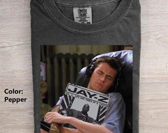 May include: A pepper-colored crew neck t-shirt featuring a graphic print of a person wearing headphones and holding a magazine titled JAY-Z LIFE AND TIMES OF S. CARTER. The shirt has a Comfort Colors brand label on the inside of the collar.