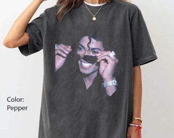 May include: A pepper-colored oversized t-shirt featuring a graphic print of a person wearing dark sunglasses, a ring, and a metallic watch. The person is smiling, showing teeth, and adjusting the sunglasses with both hands.