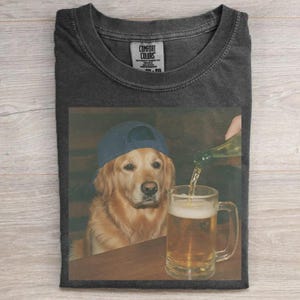 May include: Dark gray Comfort Colors t-shirt featuring a graphic of a golden retriever wearing a blue baseball cap, sitting at a bar while beer is poured into a mug. The shirt has a classic crew neck and short sleeves.