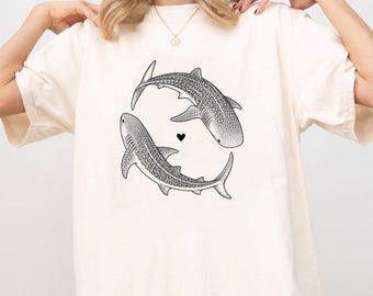 May include: A cream-colored short-sleeve t-shirt featuring a black line art graphic of two whale sharks swimming in a circular formation with a small black heart symbol positioned in the center between the two sharks on the front chest area.