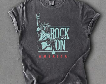 May include: A charcoal gray short-sleeve t-shirt featuring a light blue graphic of the Statue of Liberty playing an electric guitar. The text reads ROCK ON in light blue and AMERICA in red. The shirt has a crew neck and a vintage-style wash.