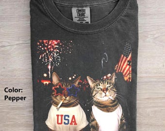 May include: A charcoal gray t-shirt with a graphic print of two tabby cats. One cat wears star-shaped red and blue glasses and a white shirt with the text USA. The other cat wears a white tank top. The background features fireworks and a US flag.