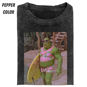 May include: Black t-shirt with a graphic of a green ogre wearing a pink floral bikini, sunglasses, and holding a yellow surfboard. The text "PEPPER COLOR" is printed in white at the top left of the shirt.