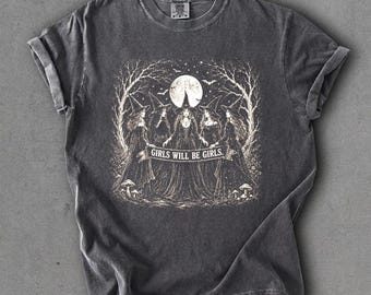 May include: A charcoal gray short-sleeve t-shirt featuring a white graphic print of five figures in witch hats standing in a circle under a full moon and bare trees. A banner across the center contains the text "GIRLS WILL BE GIRLS."