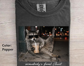 May include: A charcoal gray t-shirt featuring a graphic of a raccoon wearing black sunglasses and holding a plastic cup with a straw. The raccoon is sitting on the ground. The text "somebody's feral Aunt" is printed in white script below the image.