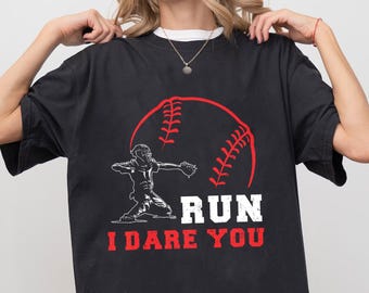 May include: Black t-shirt featuring a baseball design. A red baseball graphic is behind a white and black illustration of a baseball player. The text "RUN I DARE YOU" is printed in white and red.