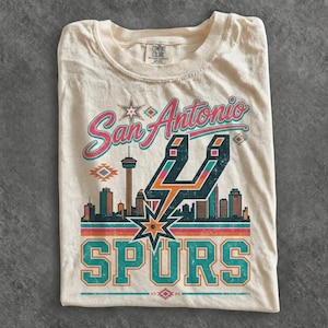 May include: A cream-colored t-shirt featuring a graphic print with the text San Antonio Spurs. The design includes a stylized city skyline, the Tower of the Americas, and the Spurs logo in teal, orange, and pink colors with geometric accents.