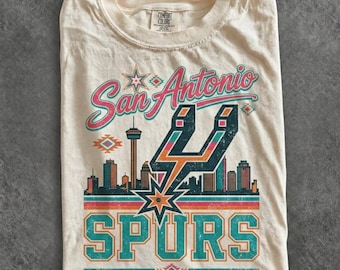 May include: A cream-colored t-shirt featuring a graphic print with the text San Antonio Spurs. The design includes a stylized city skyline, the Tower of the Americas, and the Spurs logo in teal, orange, and pink colors with geometric accents.