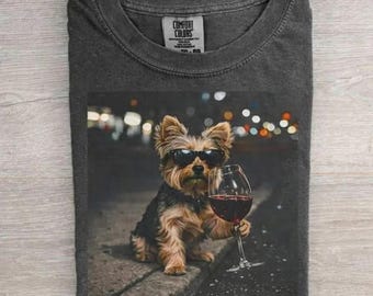 May include: Dark gray t-shirt featuring a Yorkshire Terrier wearing sunglasses and holding a glass of red wine. The dog is sitting on a ledge with blurred lights in the background. The shirt has a Comfort Colors tag.