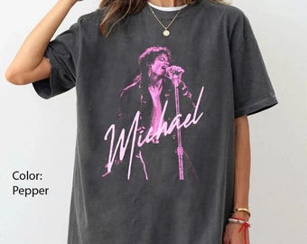May include: A pepper-colored short-sleeve t-shirt featuring a pink graphic print of a singer holding a microphone on a stand. The graphic includes the script text "Michael" in pink across the center of the chest. The shirt has a crew neckline.