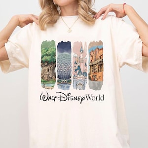May include: A cream-coloured t-shirt with a colourful graphic of Walt Disney World landmarks. The design includes images of the Tree of Life, Spaceship Earth, Cinderella Castle, and the Hollywood Tower Hotel, with "Walt Disney World" text.