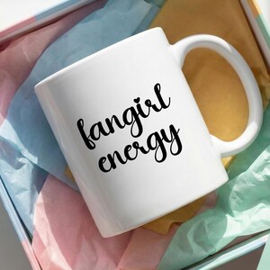 May include: White ceramic mug with the words "fangirl energy" in black script. The mug is inside a pink and blue gift box with colorful tissue paper. The mug has a large handle.