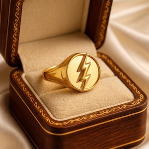 May include: A gold ring with a lightning bolt emblem inside a round face, presented in a wooden jewelry box with a cream-colored interior. The box is brown with gold trim, set against a cream-colored satin background.