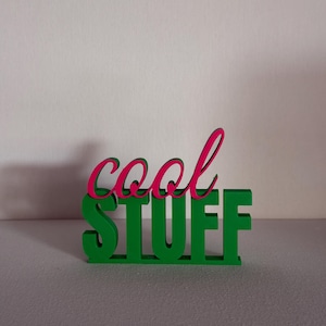 Könnte beinhalten: Ein freistehendes Schild mit dem Wort cool in einer pinkfarbenen Schreibschrift, das über dem Wort STUFF in einer fetten grünen Blockschrift angebracht ist. Das Objekt besteht aus festem Material und hat eine flache Basis für einen stabilen Stand.