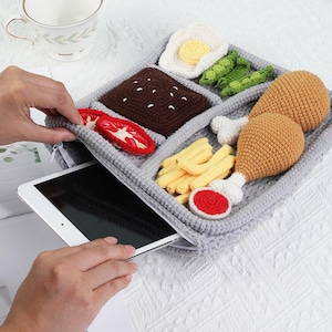 May include: A gray crocheted tablet case featuring a whimsical food design. The case is divided into sections, with crocheted representations of fried chicken, fries, a fried egg, peas, a brownie, and tomato slices. A tablet is partially visible inside.